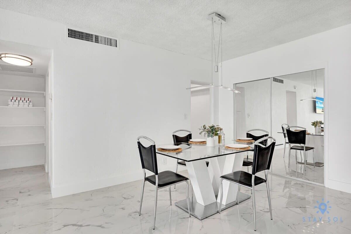Un comedor moderno con mesa de vidrio y sillas metálicas, paredes blancas y un espejo grande que refleja la habitación.