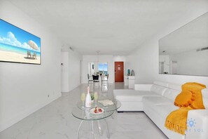 A modern living room with a glass coffee table, white sofa, and a large flat-screen TV on the wall. - Balcony-Beachfront Condo+ Gym (Hollywood)