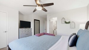 Et soveværelse med seng, ventilator, kommode, fjernsyn og en dør til et andet rum.