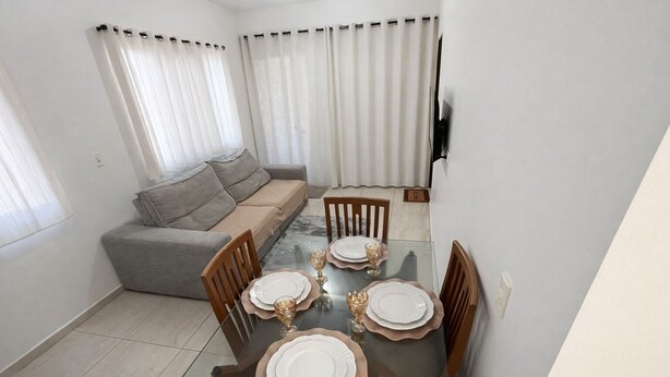 A dining area with a glass table set for four, surrounded by wooden chairs. A grey sofa is against the wall, and there are curtains on the windows. - flatportodegalinhas  (Ipojuca)