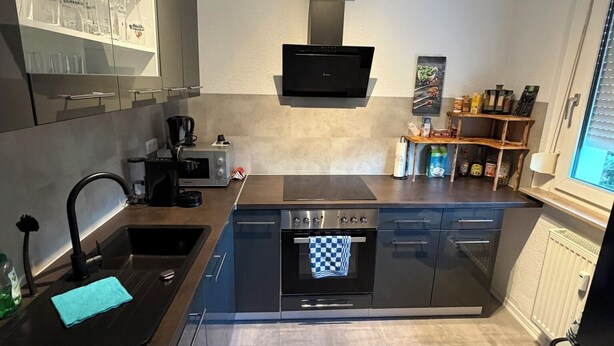 A modern kitchen with a built-in oven, a dishwasher, and a coffee machine. There is a window with blinds and a shelf with glassware.