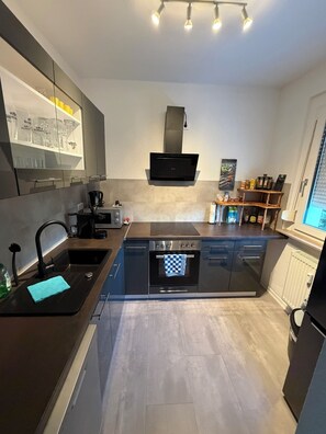 A modern kitchen with a built-in oven, a dishwasher, and a coffee machine. There is a window with blinds and a shelf with glassware. - Temporary accommodation / Fitters' apartment Zwickau-Mosel (Zwickau)