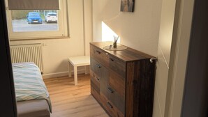 A bedroom with a bed, a wooden dresser, a radiator, a window with a view of parked cars, and a small bench.