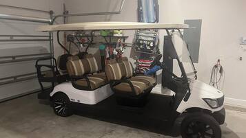 Un carrito de golf con carrocería blanca y asientos marrones, estacionado en un garaje con piso de concreto y una lámpara en el techo.