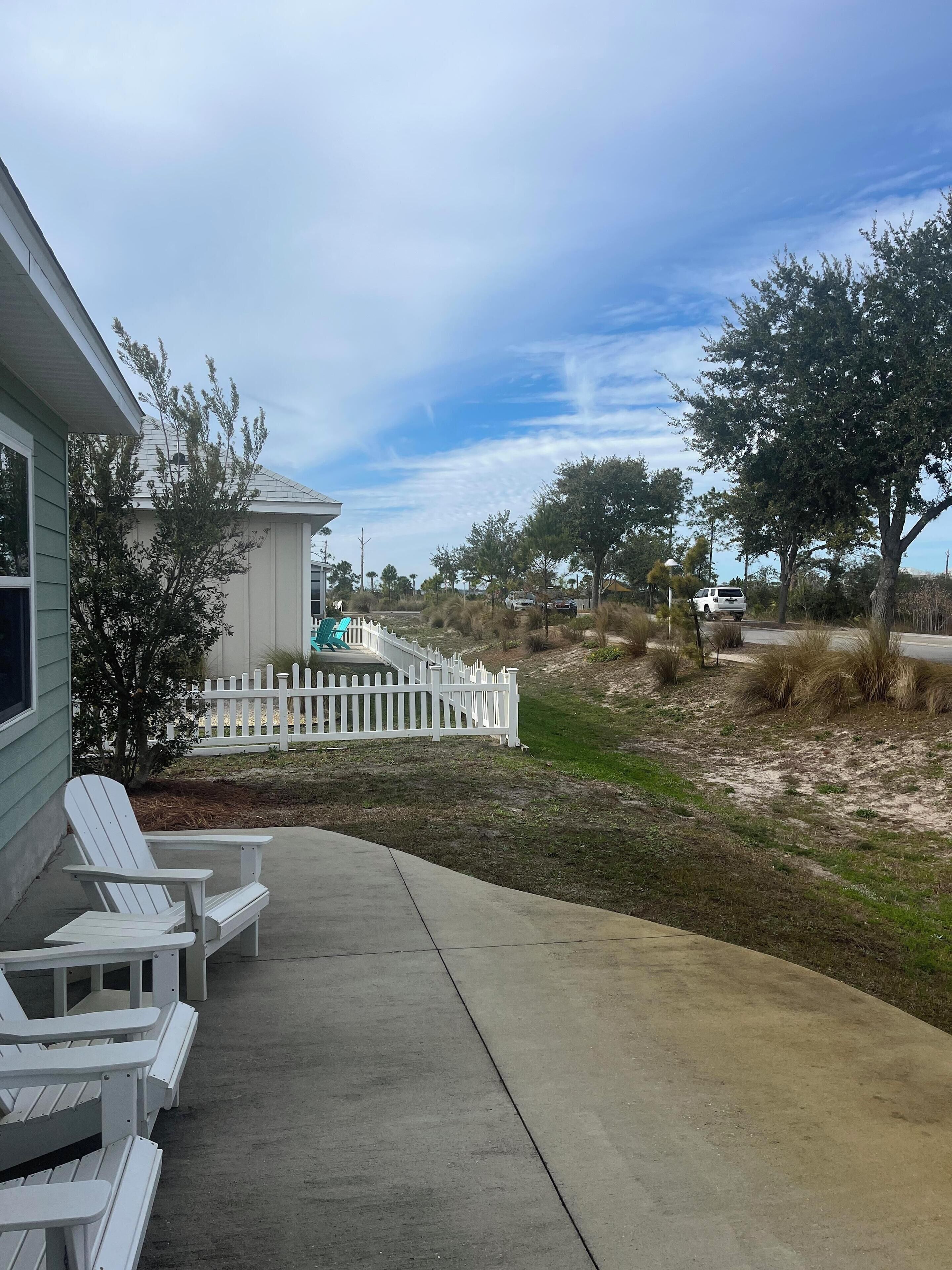 A residential property with a concrete patio, white Adirondack chairs, a white picket fence, and a grassy yard with trees.