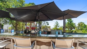 Un espace bar extérieur doté d’un comptoir en verre, de plusieurs chaises et de grands parasols offrant de l’ombre.