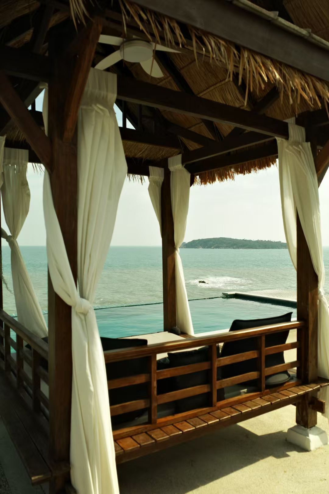 A wooden deck with a thatched roof, white curtains, and a view of the sea.