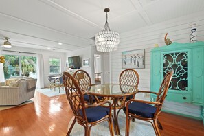 Un coin repas avec une table ronde en verre entourée de chaises en osier dotées de coussins bleus, un buffet turquoise avec des portes en verre et un lustre hexagonal suspendu.