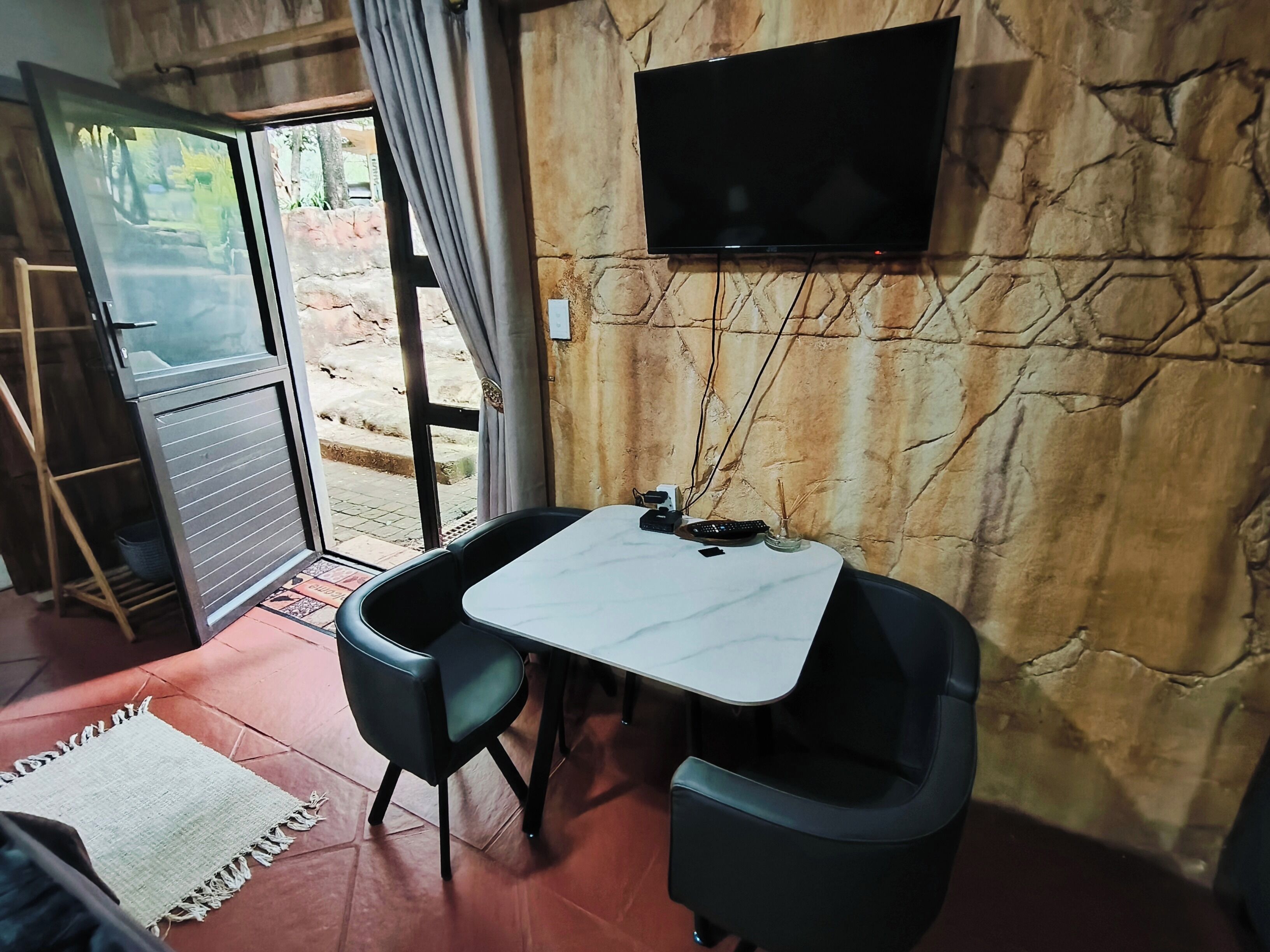A small room with a stone wall, a flat-screen TV mounted above a table with two chairs, a remote control, and a small decorative item on the table.