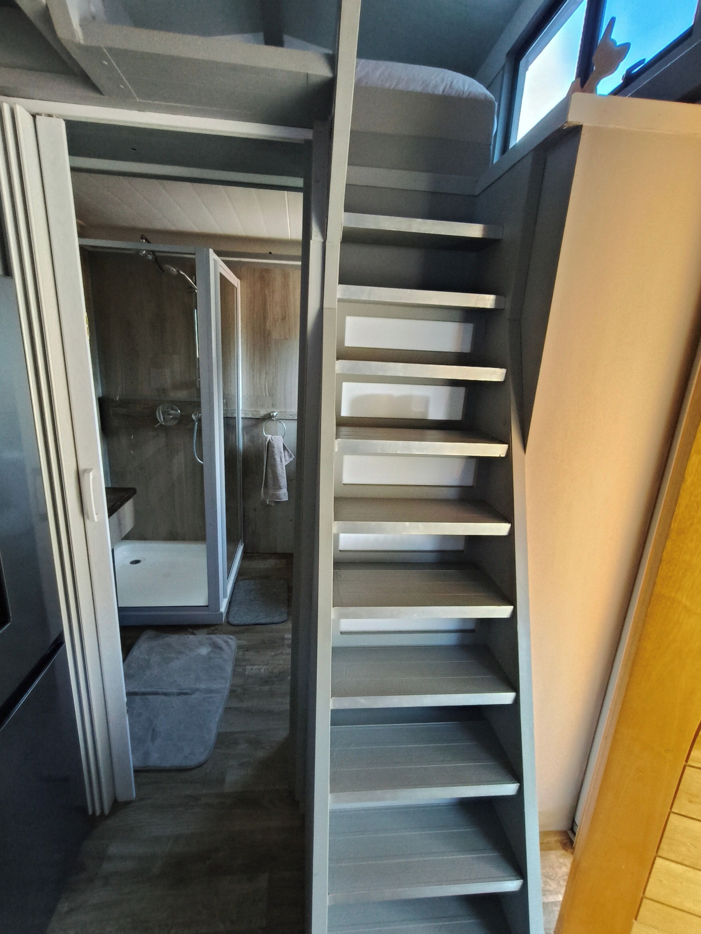 A narrow hallway with a staircase leading to a bedroom. The room has a shower, a towel hanging on the wall, and a window with a view of the sky.