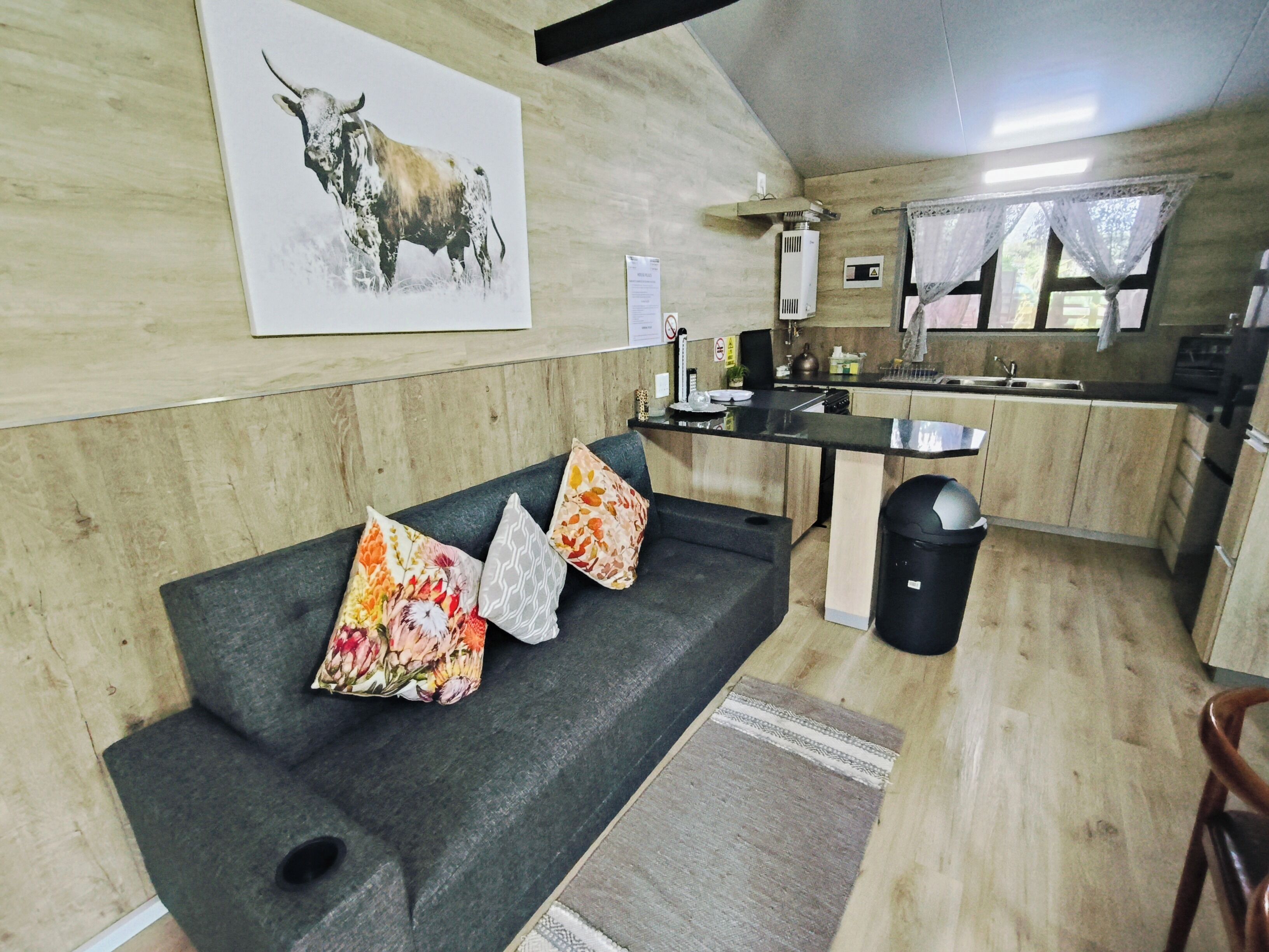 A modern kitchen with a black countertop, wooden cabinets, and a sink. There is a black sofa with floral cushions and a rug on the floor.