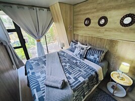 Un dormitorio con cama, cortinas, una lámpara de noche y vistas a la vegetación.