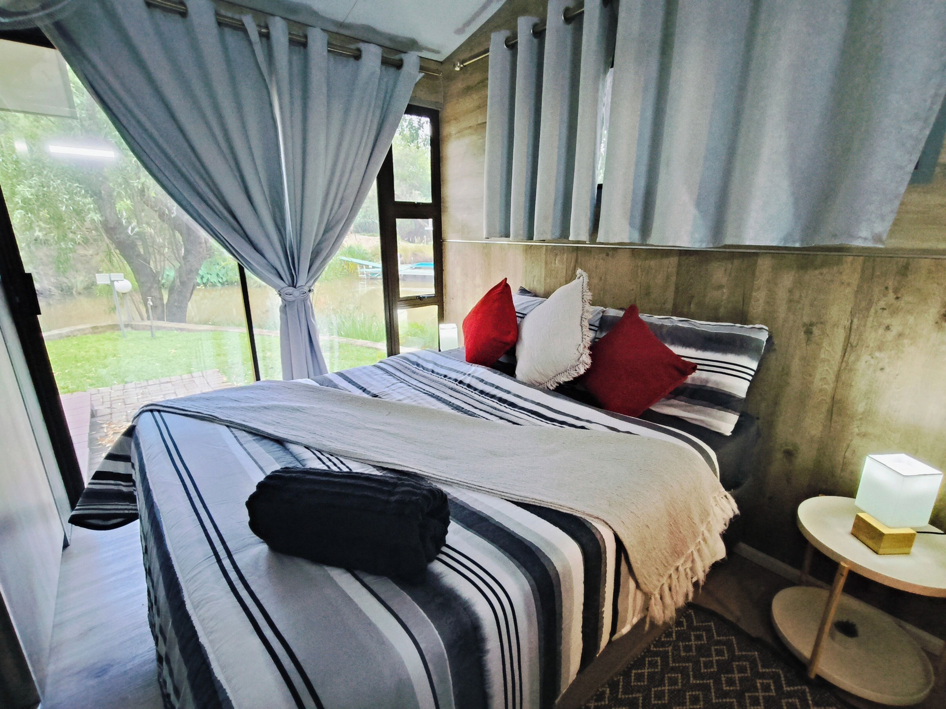 A bed with striped bedding and red pillows, a bedside table with a lamp, and a view of greenery outside.