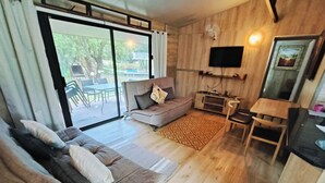 A modern living room with a sofa, a TV mounted on the wall, and a dining area visible through a glass door. - Lost Valley Farm Lodge (Rustenburg)
