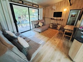 Una sala de estar moderna con un sofá, un televisor montado en la pared y un comedor visible a través de un ventanal.