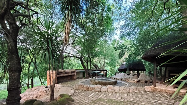 A paved courtyard with stone benches and a central water feature, surrounded by trees and a thatched-roof structure. - Lost Valley Farm Lodge (Rustenburg)