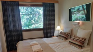 A hotel room with two beds, a TV mounted on the wall, a window with curtains, and a bedside lamp.