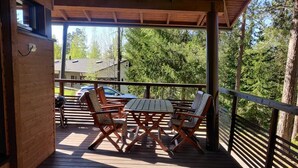 A covered outdoor deck with a wooden table and chairs, surrounded by a railing, overlooking a forested area.