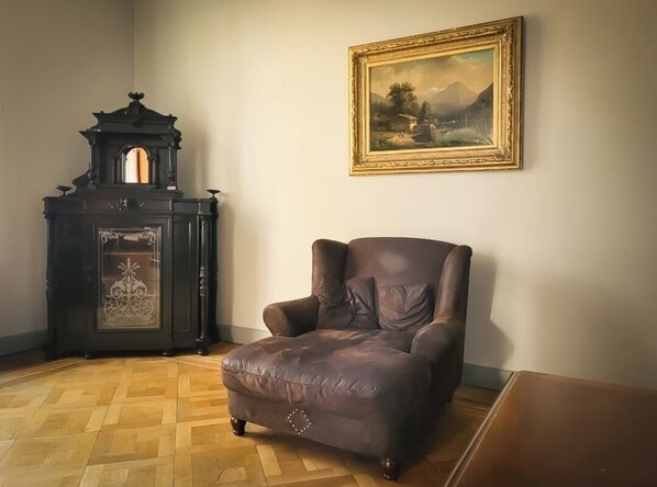 A room with a brown leather armchair, a dark wooden cabinet with intricate designs, and a framed landscape painting on the wall.