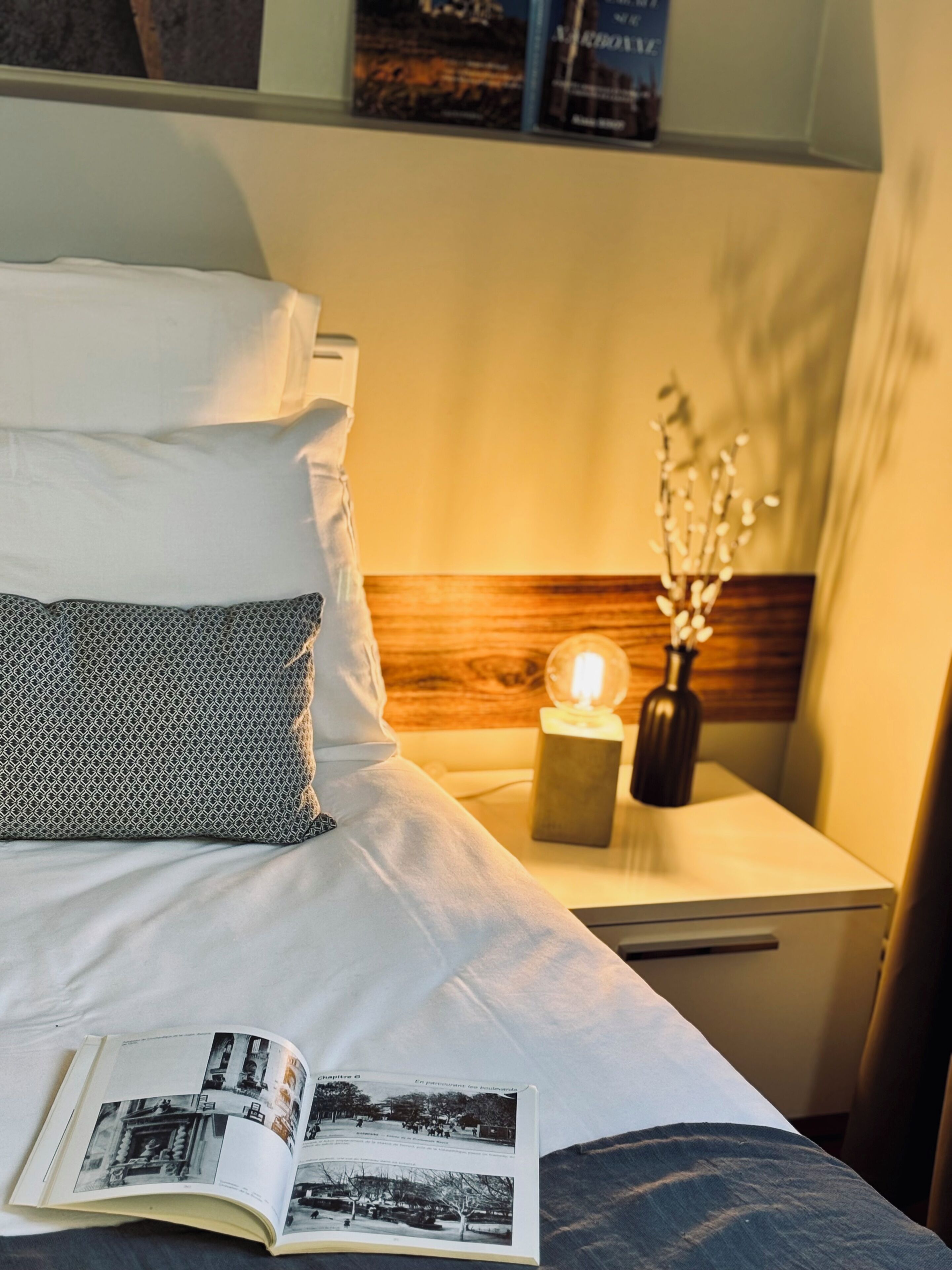 A neatly made bed with a book open on it, a bedside table with a lamp and a vase of flowers, and a wooden headboard.