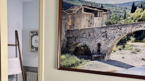 Habitació amb una fotografia en marc d'un pont de pedra sobre un riu, un radiador, una escala i un interruptor de llum a la paret.