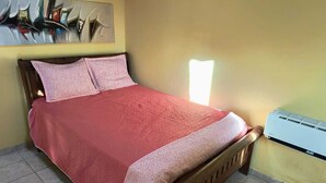 Een bed met een roze dekbedovertrek en passende kussens, een houten hoofdbord en een aan de muur bevestigde airconditioner.