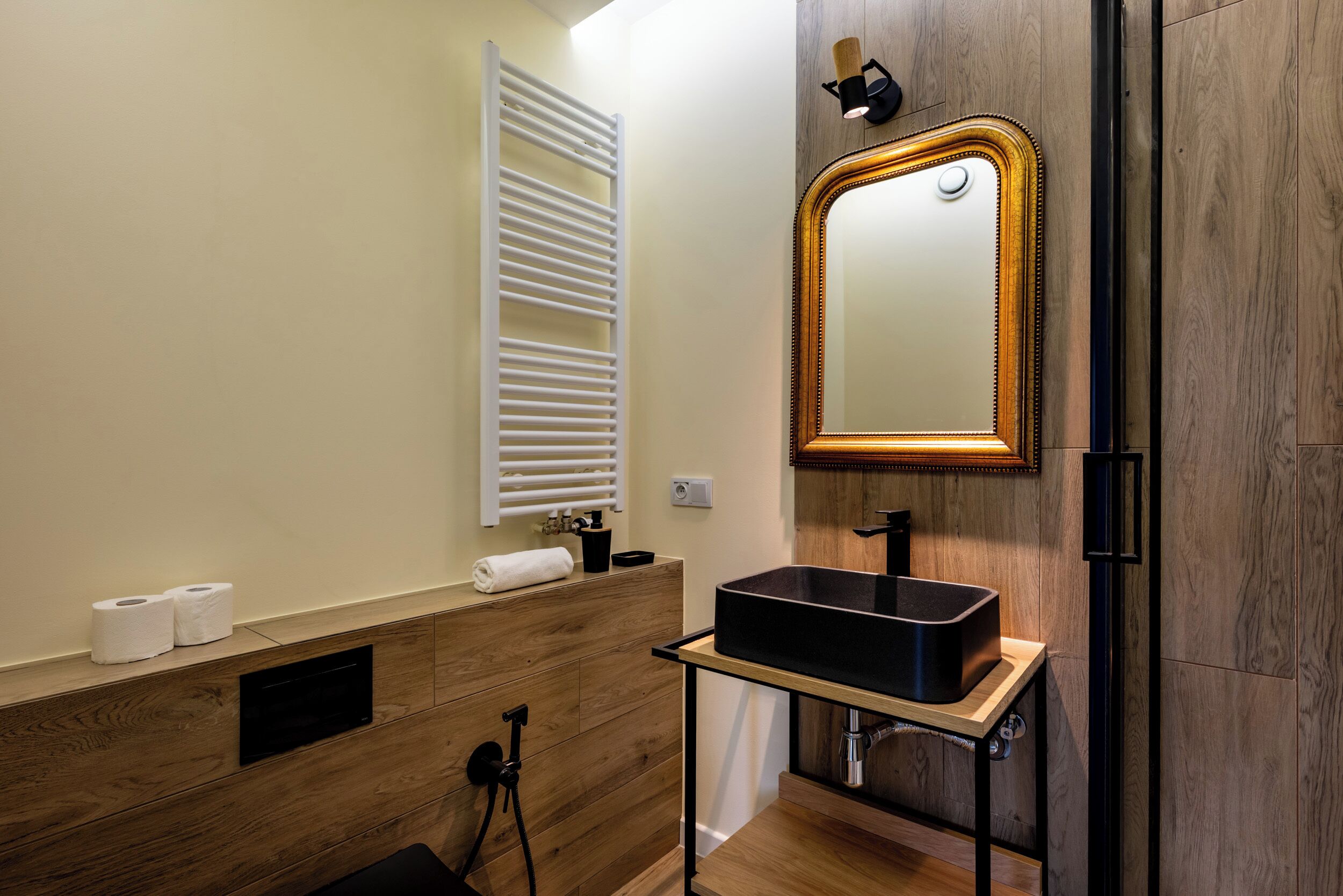 Ein modernes Badezimmer mit einem Holzbadezimmer-Möbel, einem schwarzen Waschbecken, einem Spiegel mit goldener Rahmung und einem Handtuchhalter.
