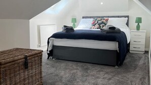 Un dormitorio con techo inclinado, una cama con cabecera oscura, una mesita de noche con una lámpara verde y una cesta de mimbre.