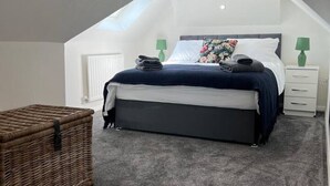 A bedroom with a sloped ceiling, a bed with a dark headboard, two bedside tables with lamps, and a wicker chest of drawers. - Gatwick 2 bedroom Apartment (Grasmere)
