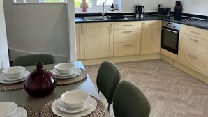 A modern kitchen with light wood cabinets, a black countertop, and a built-in oven. There is a dining area with a table set for four, including plates, bowls, and a decorative vase. - Gatwick 2 bedroom Apartment (Grasmere)