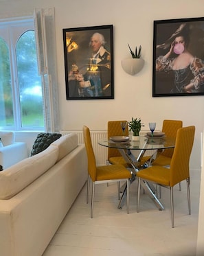 A dining area with a glass table and yellow chairs, a white sofa, and two framed artworks on the wall. - Spacious 2 bed apartment in Addlestone (Surrey)