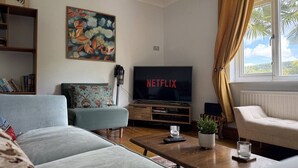 Olohuoneessa on televisio, jossa näkyy teksti »NETFLIX», sohva, lasilla varustettu sohvapöytä ja ikkuna, jossa on verhot.