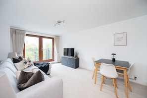 A modern living room with a sofa, a dining table with chairs, a TV, and a balcony with curtains. - 2 Bedroom Flat Free Parking Close to Gatwick (West Sussex)