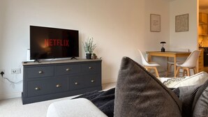 Ein modernes Wohnzimmer mit einem Flachbildfernseher auf einer dunklen Kommode, einer Essecke mit Holztisch und -stühlen sowie einer Küche im Hintergrund.