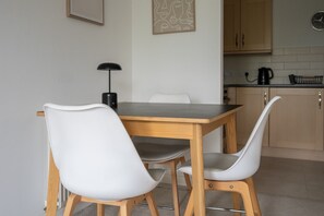 A dining area with a wooden table and white chairs, a black lamp, and a kitchen in the background. - 2 Bedroom Flat Free Parking Close to Gatwick (West Sussex)