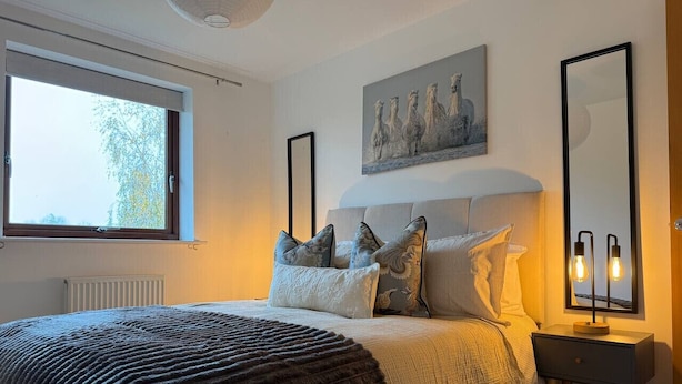 Ein Schlafzimmer mit einem großen Bett, einem Nachttisch, einer Wandleuchte und einem Fenster mit Blick auf Bäume.