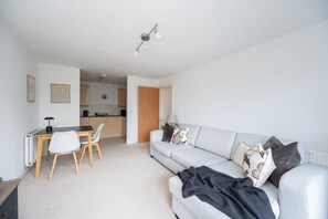 Ein modernes Wohnzimmer mit einem grauen Sofa, einem Esstisch mit Stühlen und einer Küche im Hintergrund.