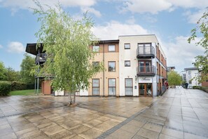 A multi-story building with a storefront on the ground floor, a tree in the foreground, and a paved walkway. - 2 Bedroom Flat Free Parking Close to Gatwick (West Sussex)