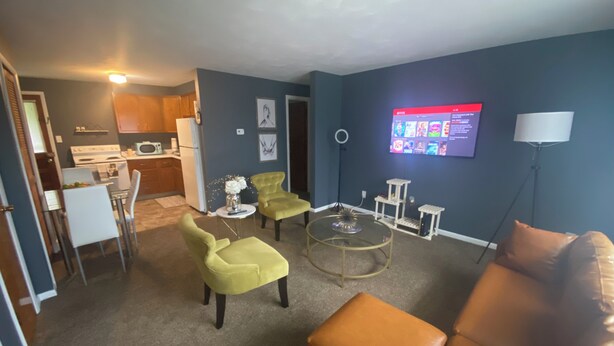 Uma sala de estar com um sofá de couro marrom, duas poltronas amarelas, uma mesa de centro de vidro, uma TV fixada na parede e uma área de cozinha com armários de madeira e um micro-ondas.