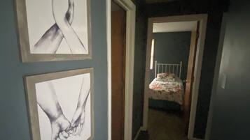 Un couloir orné de deux œuvres d’art encadrées, une porte donnant sur une chambre à coucher, et un lit recouvert d’une couvre-lit à motifs floraux.