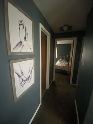A hallway with two framed artworks on the wall, a door leading to a bedroom, and a bed with a floral bedspread.