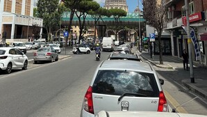 A busy urban street with parked cars, a prominent dome structure in the background, and multi-story buildings with balconies. - Vatican YH (Roma)
