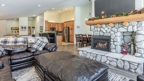 A modern living room with a stone fireplace, leather sofa, and a kitchen in the background. - Fairway Front Luxury with Sweeping Mountain Views | 4BR (Mammoth Lakes)