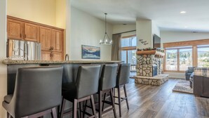 A modern kitchen with a stone fireplace, bar seating, and large windows offering views of the outdoors. - Fairway Front Luxury with Sweeping Mountain Views | 4BR (Mammoth Lakes)