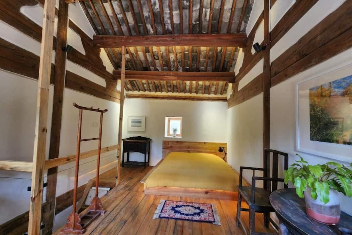 A rustic room with a wooden ceiling, a bed with a yellow bedspread, a small table, a chair, and a potted plant.