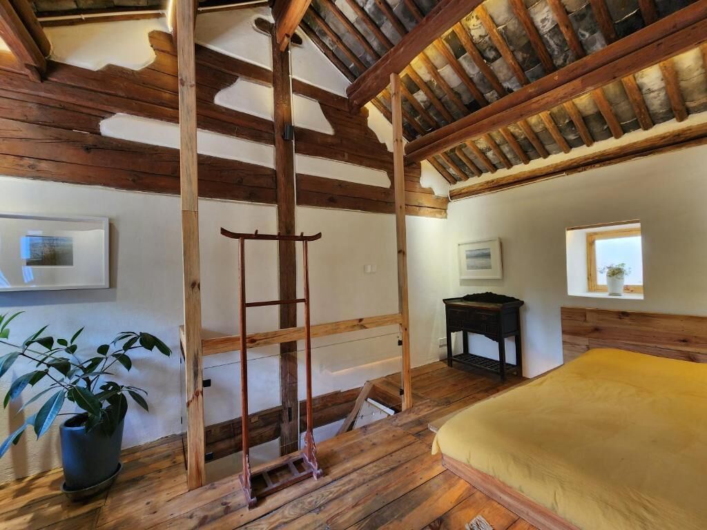 Quarto rústico com vigas de madeira aparente, escada, cama com colcha amarela, mesinha e planta em vaso.