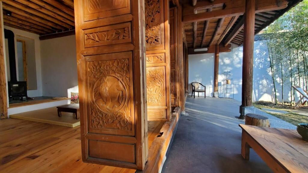 Un interior de madera con un banco y una mesa, una puerta de madera con adornos tallados, y vistas a un jardín exterior.
