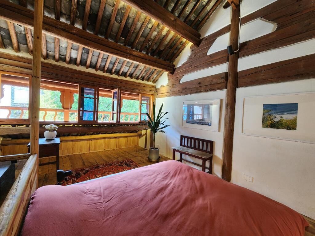 A bedroom with a wooden ceiling, large windows, a bed, a bench, and a potted plant.