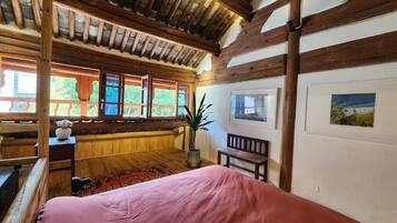 A bedroom with a wooden ceiling, large windows, a bed, a bench, and a potted plant.