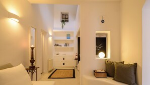 A modern hotel room with a bed, a seating area with cushions, a small table, a mirror, a wall-mounted light, and a shelf with decorative items. - Coat Of Arms Mansion (Santorini)